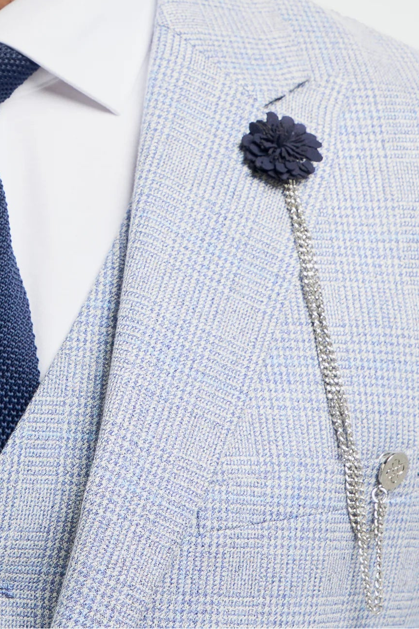 Muž ve světle šedém obleku s tmavě modrou kravatou a květinovou sponou na bílém pozadí